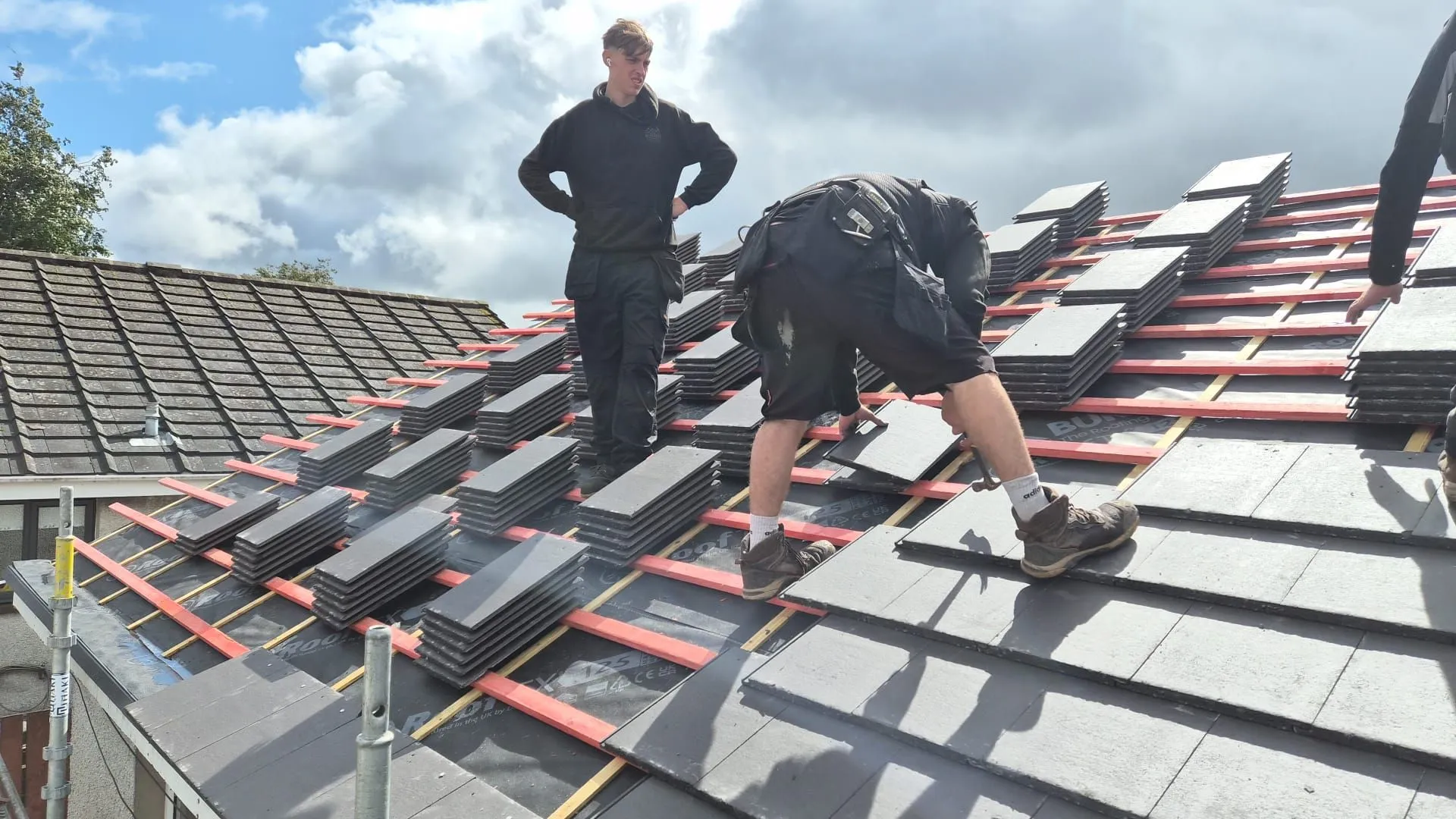 Roofer nailing down new slate grey tiles with stacks of tiles loaded out across the roof